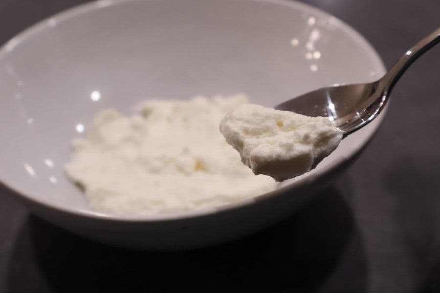 Spoonful of thick, white hung curd held above a white bowl filled with creamy curds