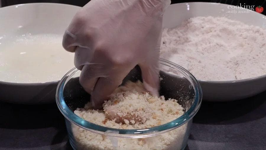 Hand pressing chicken nugget into breadcrumbs in glass bowl during breading process.