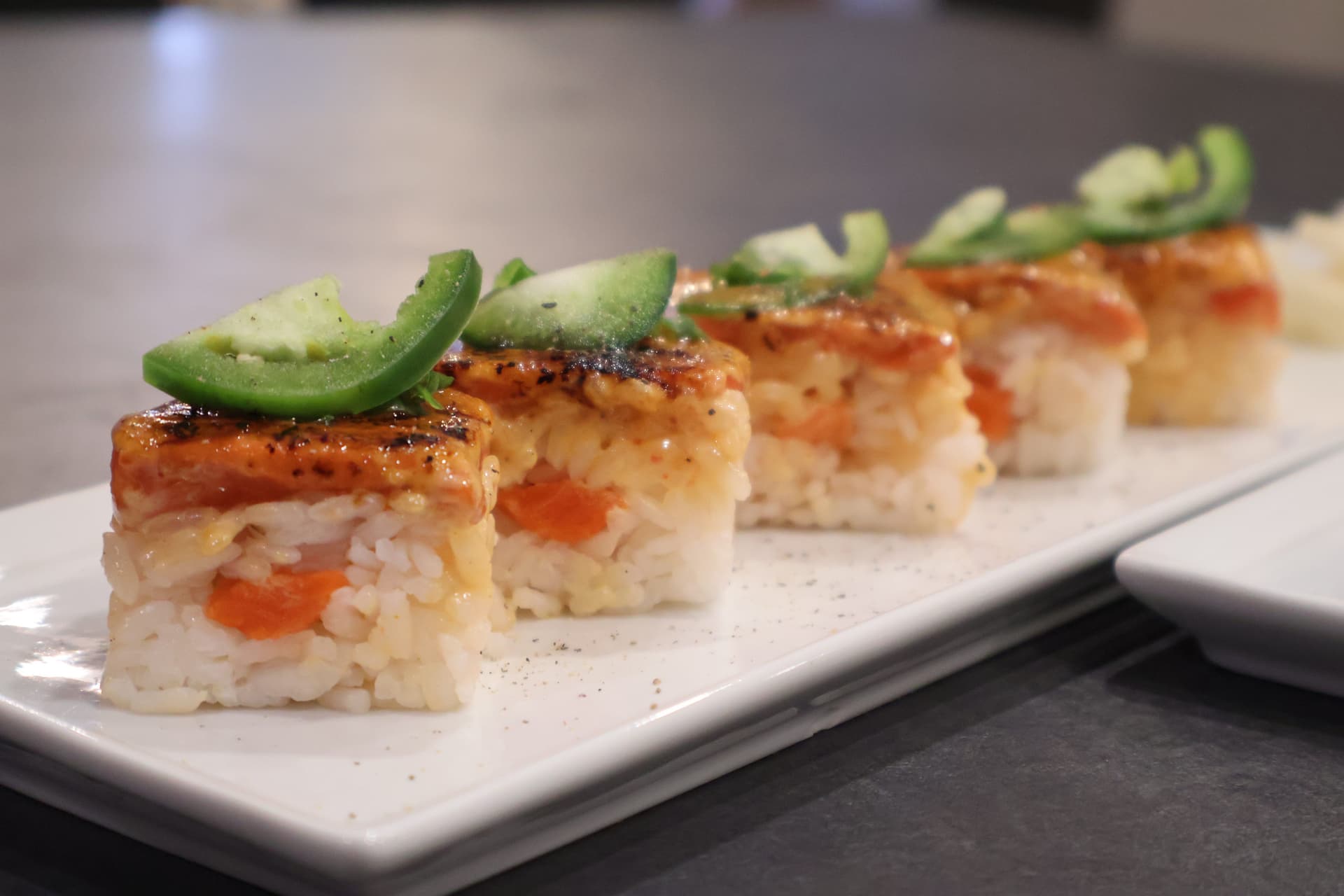 Oshizushi pressed sushi pieces topped with glazed topping and cucumber slices, arranged on a white rectangular plate.
