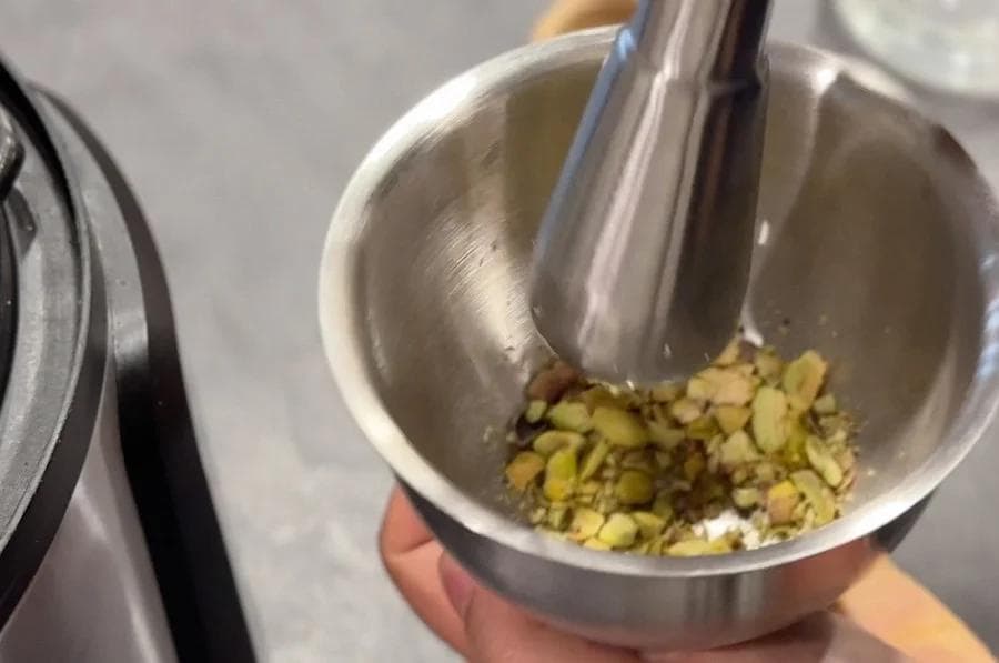 Crushing green cardamom pods in a mortar and pestle over a stainless steel bowl.