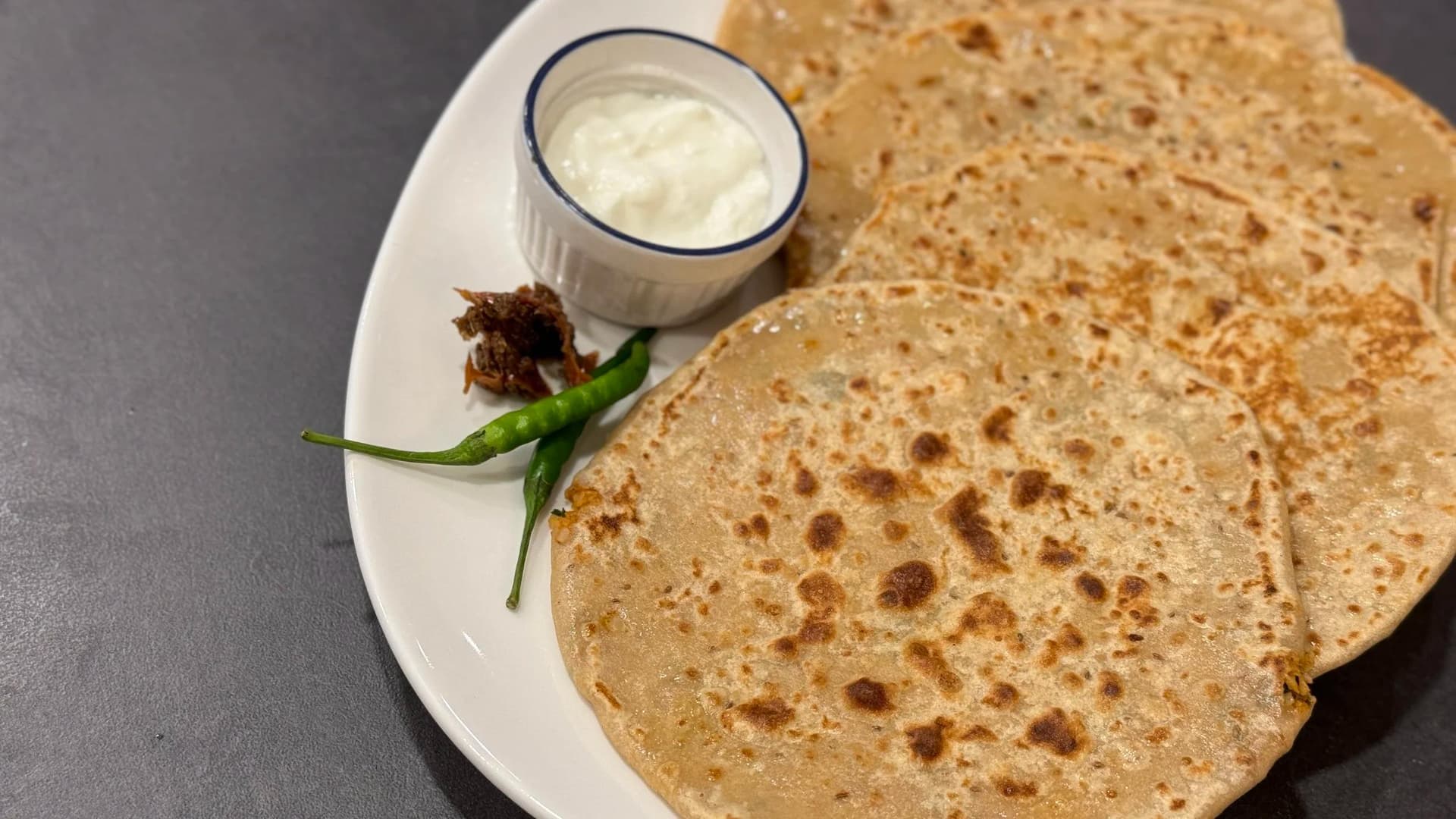 Two cooked gobi parathas on a white plate with a small bowl of yogurt and green chili on the side.