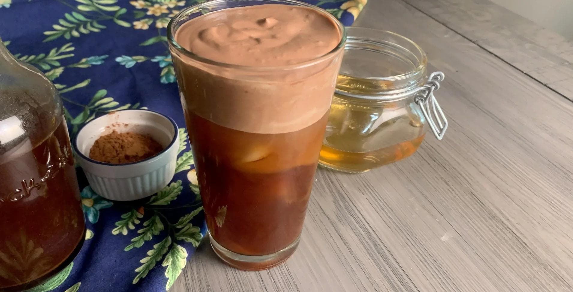 Tall glass of chocolate cold brew topped with thick chocolate foam, served on a blue floral napkin with honey and cocoa powder nearby.