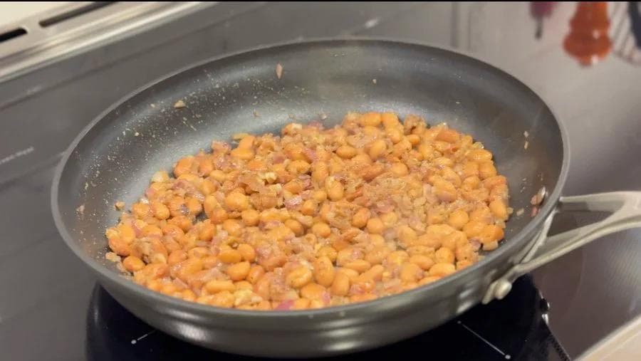 Cooked beans sautéing in a pan with spices and aromatics on stovetop.