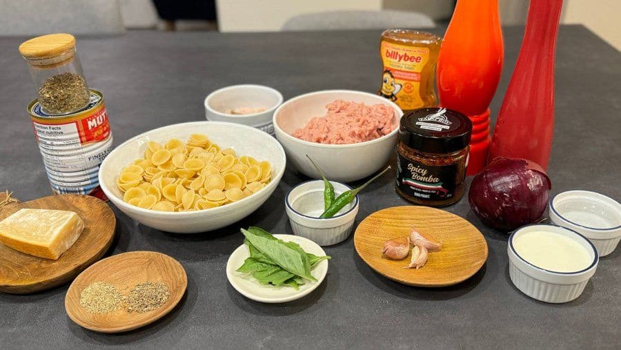 Ingredients for spicy orecchiette with Italian chicken sausage arranged on a dark countertop, including pasta, ground chicken, fresh basil, garlic, and condiments.