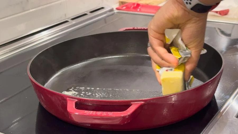 Hand buttering a red cast iron pan on a stovetop for toasting burger buns.
