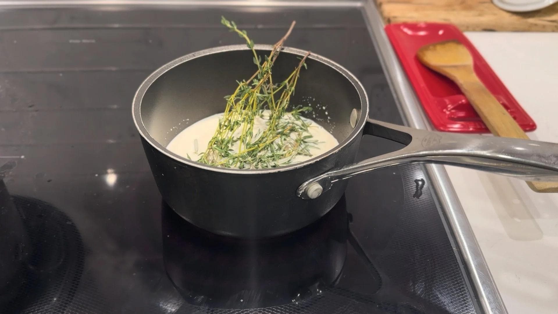 Fresh thyme and rosemary sprigs infusing in cream in a black saucepan on a stovetop.