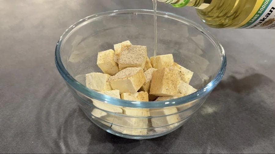Oil being drizzled over cubed tofu coated with seasonings in a glass bowl.