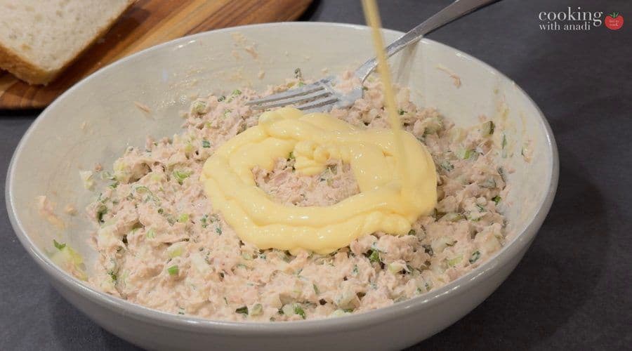 Mayonnaise being folded into a bowl of ground tuna salad mixture with herbs and pickles.