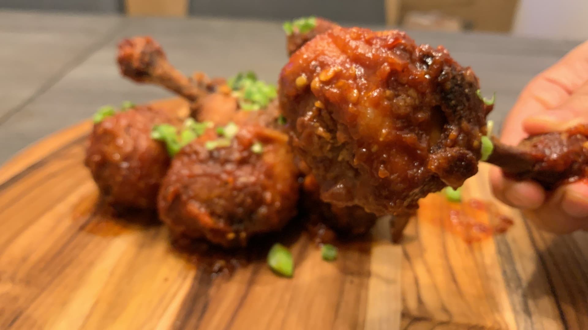 Indo-Chinese chicken lollipop drumsticks glazed in reddish-brown sauce, garnished with green onions on a wooden board.
