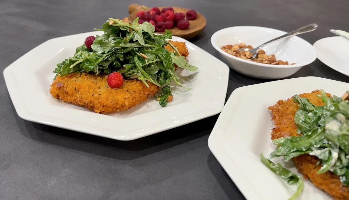 Panko-crusted chicken schnitzel topped with arugula and fresh raspberries, plated on white dishes with garnishes and nuts in bowls.