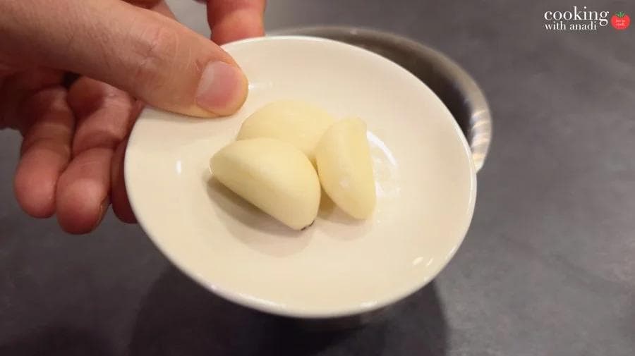 Three peeled garlic cloves in a small white bowl, held by hand, ready to be crushed into paste.
