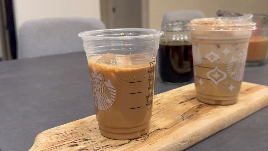 Three iced chai lattes in Starbucks cups on a wooden board, showing the layered drink with foam topping and spice garnish.