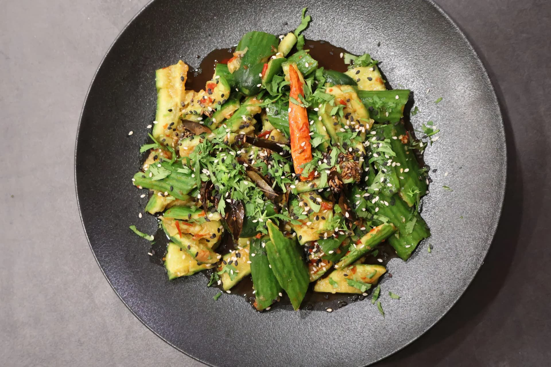 Indian fusion smashed cucumber salad with green beans, sesame seeds, and chili flakes in a dark bowl.