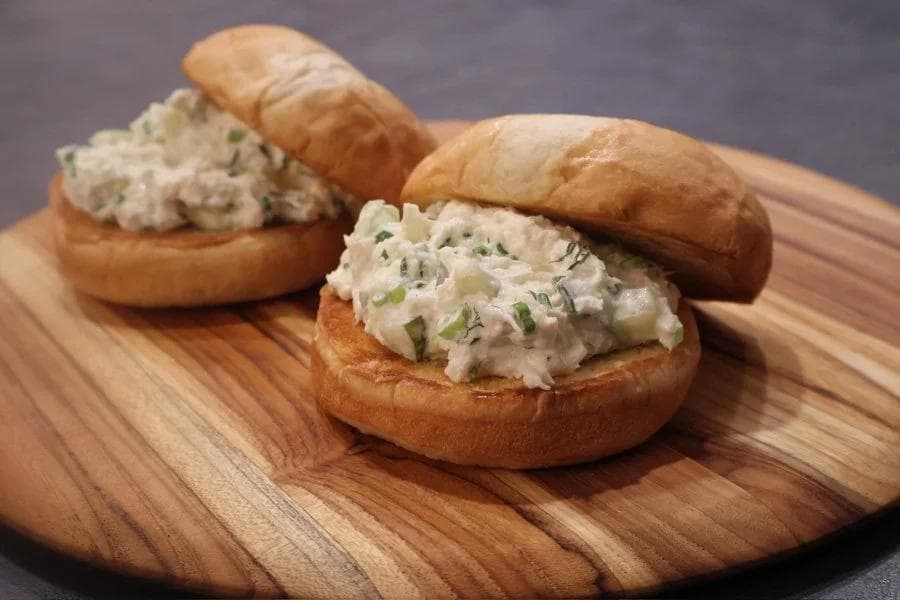 Two crab salad sandwiches on toasted buns with green onion garnish on a wooden serving board.