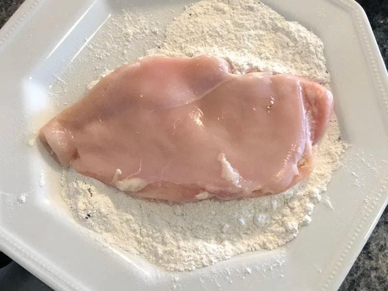 Raw chicken breast being coated in seasoned flour on a white plate for breading.