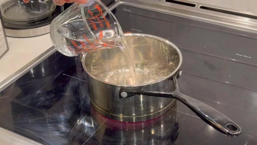 Pouring water from a measuring cup into a saucepan containing dissolved brown sugar on a stovetop.