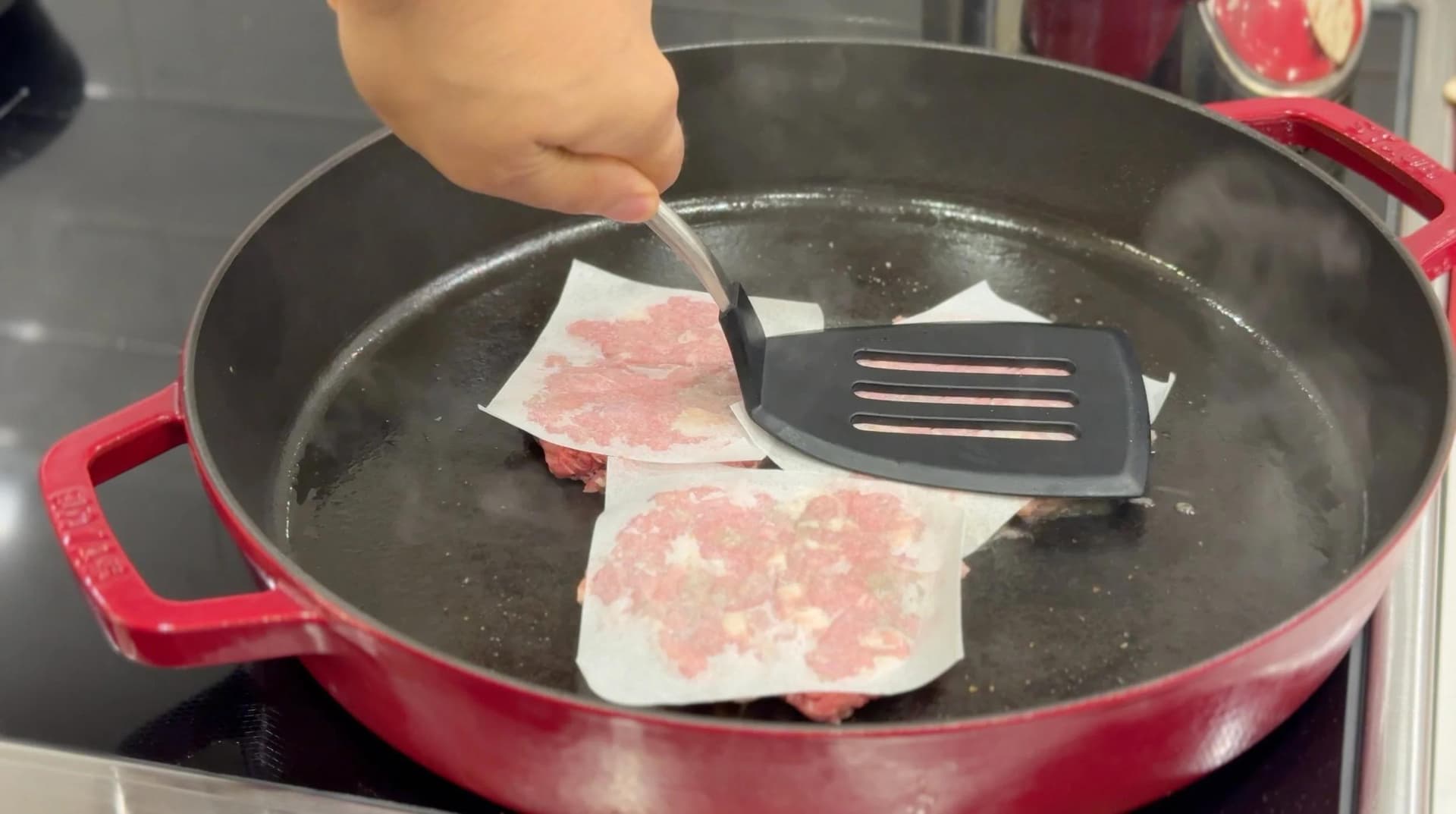 Thin sliced meat being placed on a hot red cast iron pan with a spatula during slider preparation.