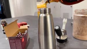 Liquid being poured from a bottle into a stainless steel whipped cream dispenser on a counter.