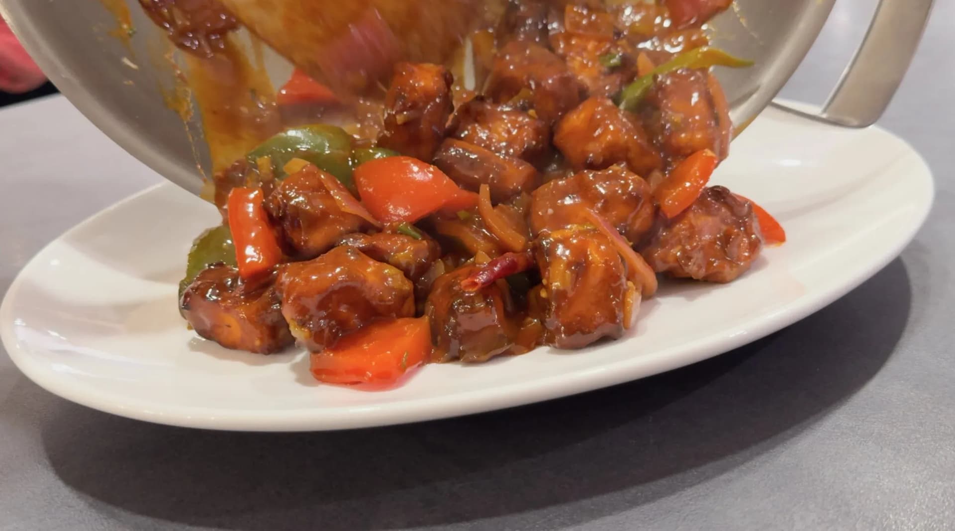 Chilli paneer being poured from a wok onto a white plate, coated in glossy red sauce with bell peppers and carrots.