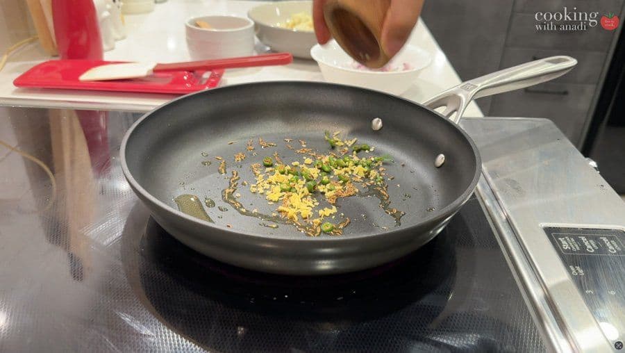 Sautéing spices including hing, cumin seeds, green chiles and ginger in oil in a dark skillet on stovetop.