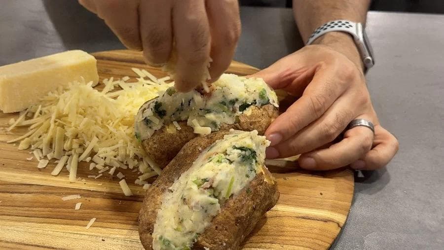 Hands spooning colcannon filling into halved potato shells topped with melted cheddar cheese on a wooden board.