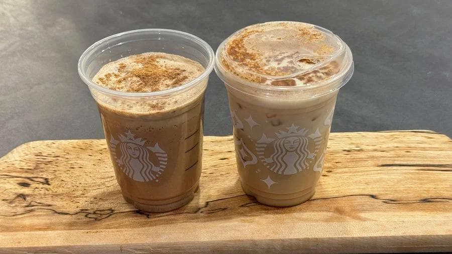 Two iced gingerbread chai lattes in Starbucks cups topped with cinnamon, displayed on a wooden board.