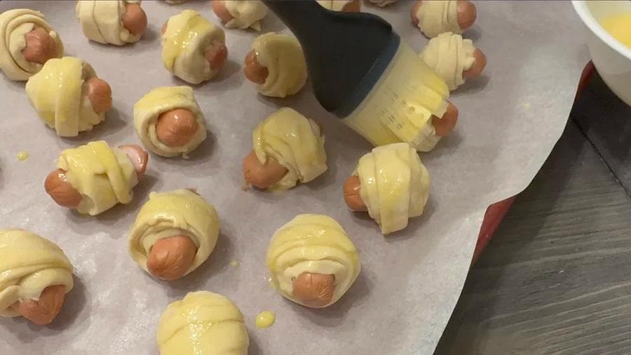 Unbaked pigs in a blanket wrapped in crescent roll dough with egg wash, arranged on parchment paper ready for air frying.