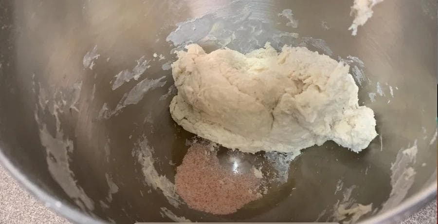 Pizza dough being kneaded in a mixer bowl with salt and olive oil added, showing soft, pliable dough texture.