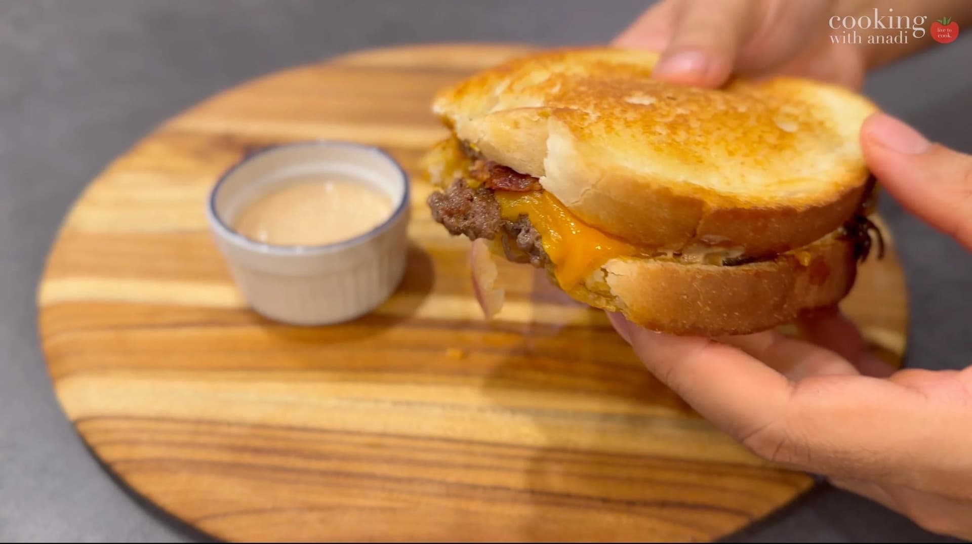 Golden-brown smash burger patty melt with melted cheese and bacon, held in hands over wooden board with sauce.
