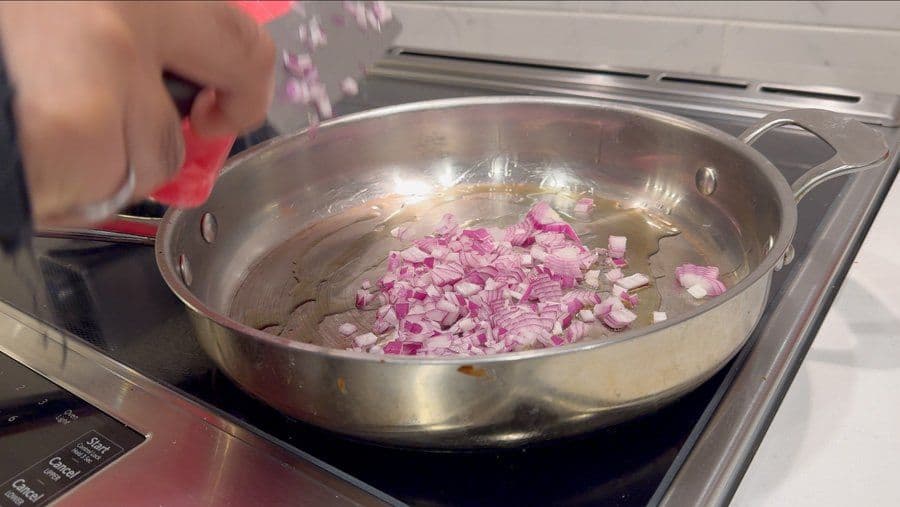 Diced red onions sautéing in olive oil in a large stainless steel pan on the stovetop.