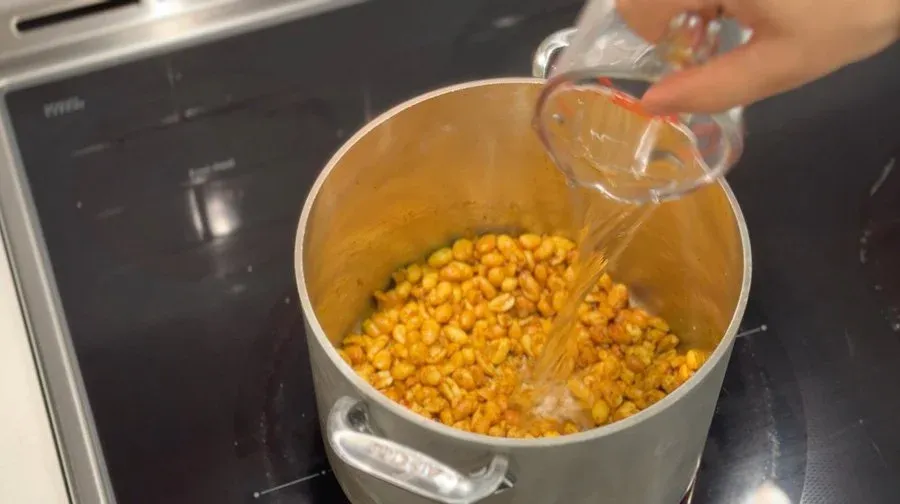 Water being poured into a pot of roasted corn kernels mixed with spices for peanut sauce.