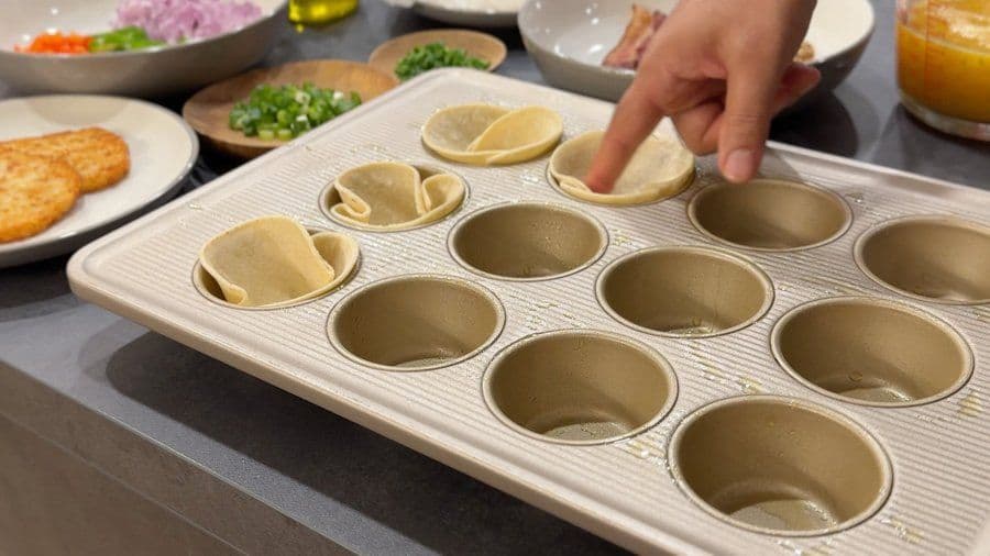 Hand placing folded tortilla into muffin tin cups lined with partially pressed tortillas for egg cups.