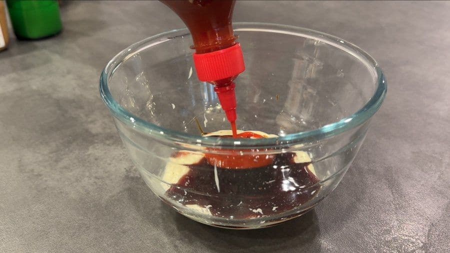 Red squeeze bottle dispensing dark umami sauce into a glass bowl containing liquid ingredients.