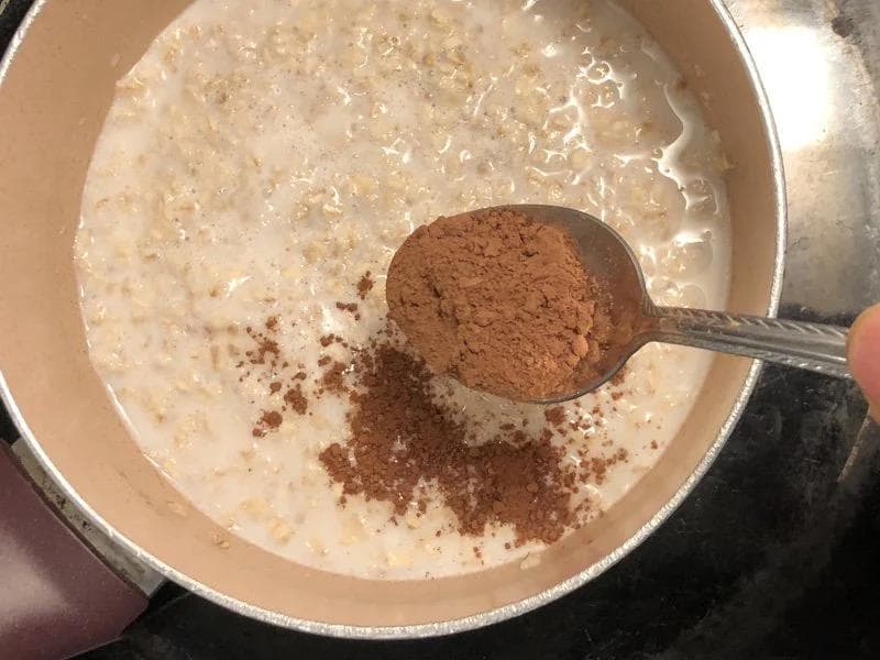 Cocoa powder being added to creamy oatmeal mixture with a spoon in a pot.