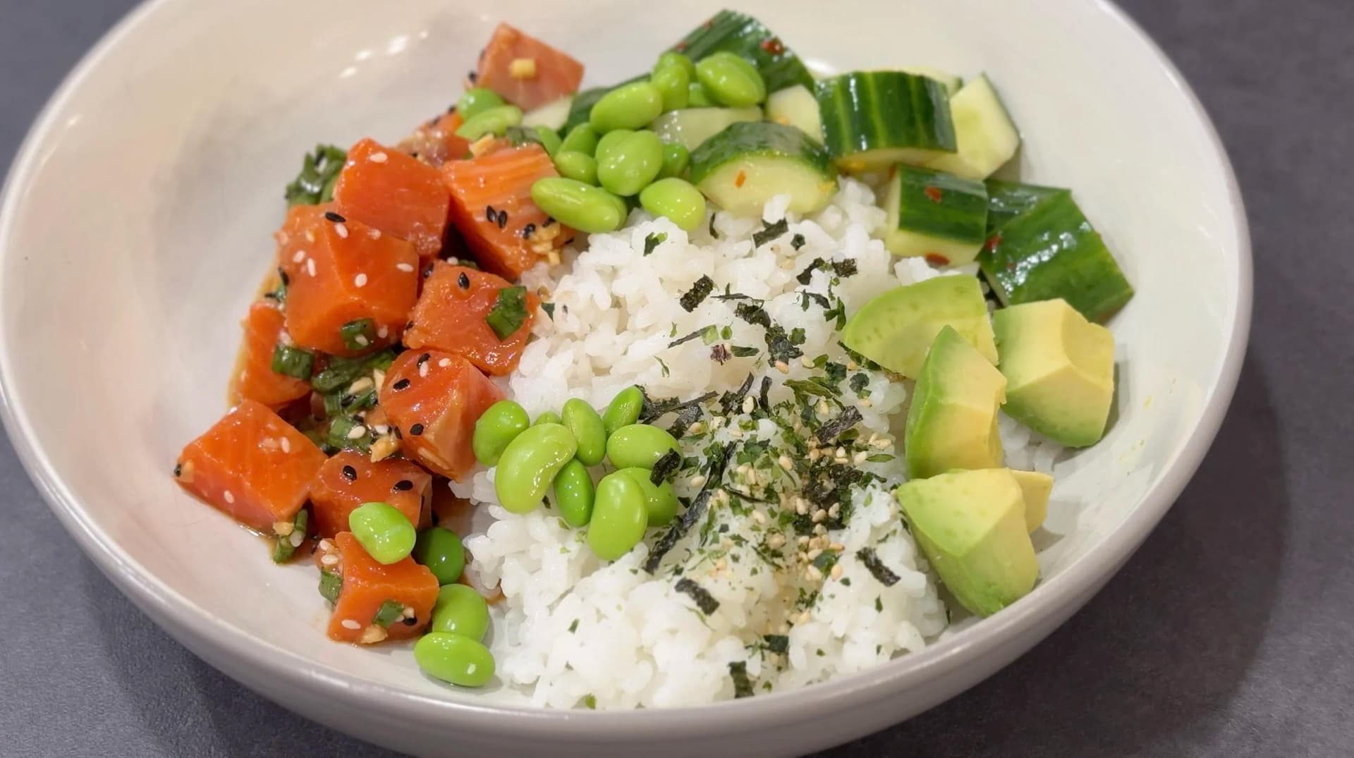 Assembled poke bowl with white rice topped with diced salmon, edamame, avocado, cucumber, nori, and furikake garnish in a white bowl.