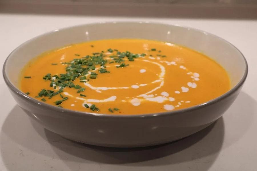 Creamy orange carrot and ginger soup garnished with fresh chives and coconut milk drizzle in a white bowl.