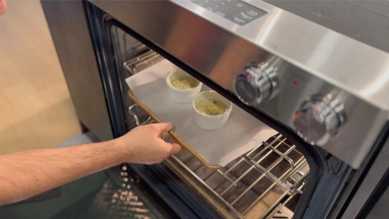 Hand pulling out a baking sheet with two ramekins of baked cheese egg soufflés from the oven.