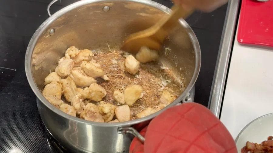 Flour-coated chicken pieces browning in bacon fat in a large pot with a wooden spoon stirring.