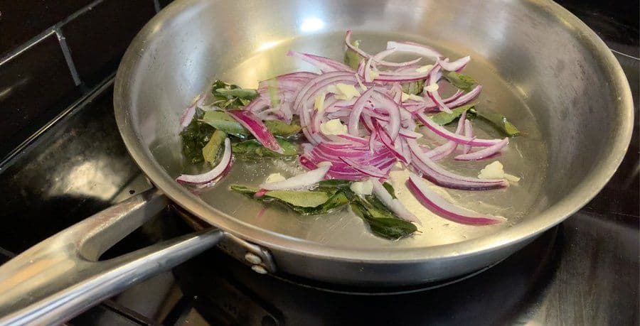 Sliced red onions and curry leaves sautéing in a stainless steel wok on a stovetop.
