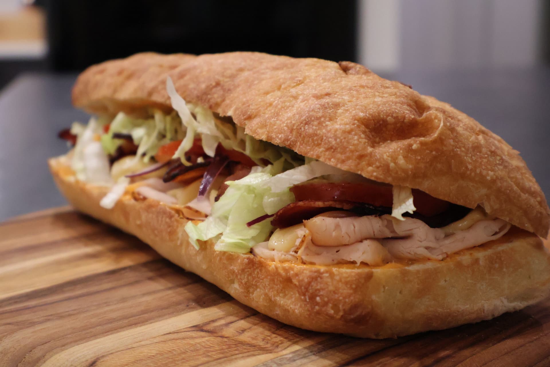 Baked deli chicken sandwich on a wooden board with melted cheese, lettuce, and tomato on crusty bread.