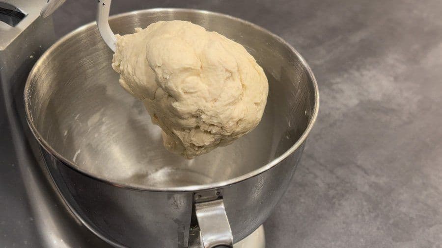 Pizza dough ball attached to dough hook in stainless steel mixing bowl after kneading with olive oil.