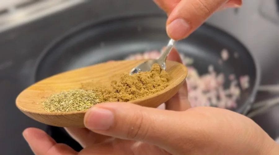 Hand holding wooden spoon with ground cumin being added to pan of diced onions for refried beans.