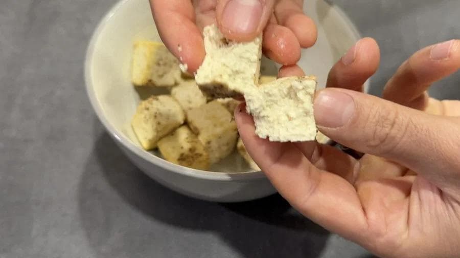 Hand holding crispy air fryer tofu cube with cornstarch coating over bowl of golden tofu pieces.
