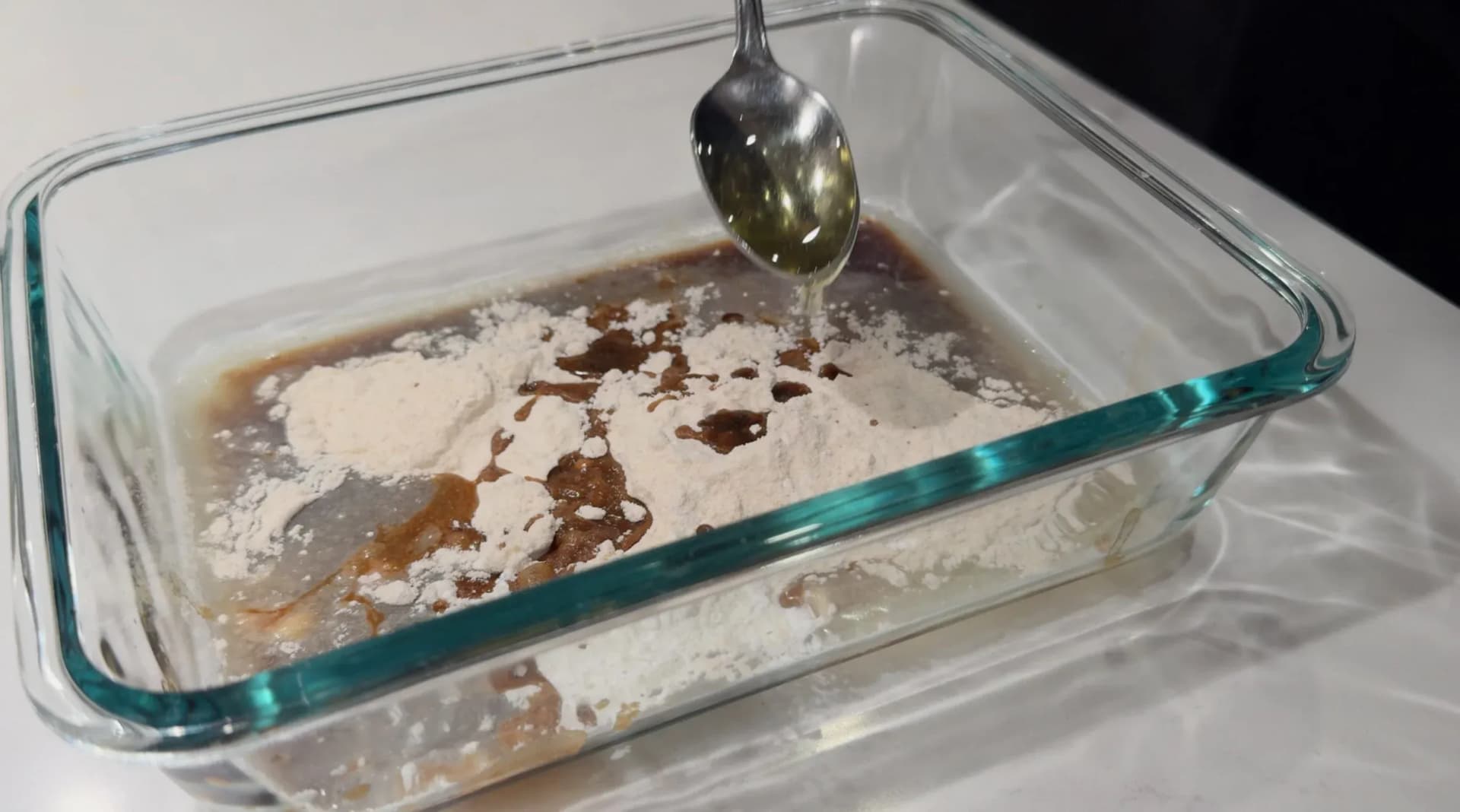 Dry batter ingredients with flour and dark liquid in a glass baking dish, with a spoon pouring liquid from above.