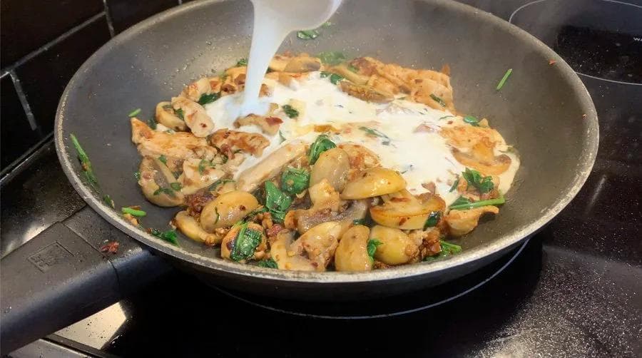 Creamy sauce being poured over sautéed mushrooms and spinach in a pan with fresh herbs.