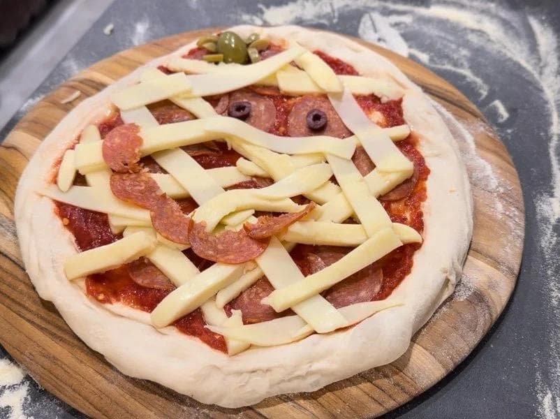 Unbaked mummy pizza with pepperoni and cheese lattice strips on a wooden board before baking.