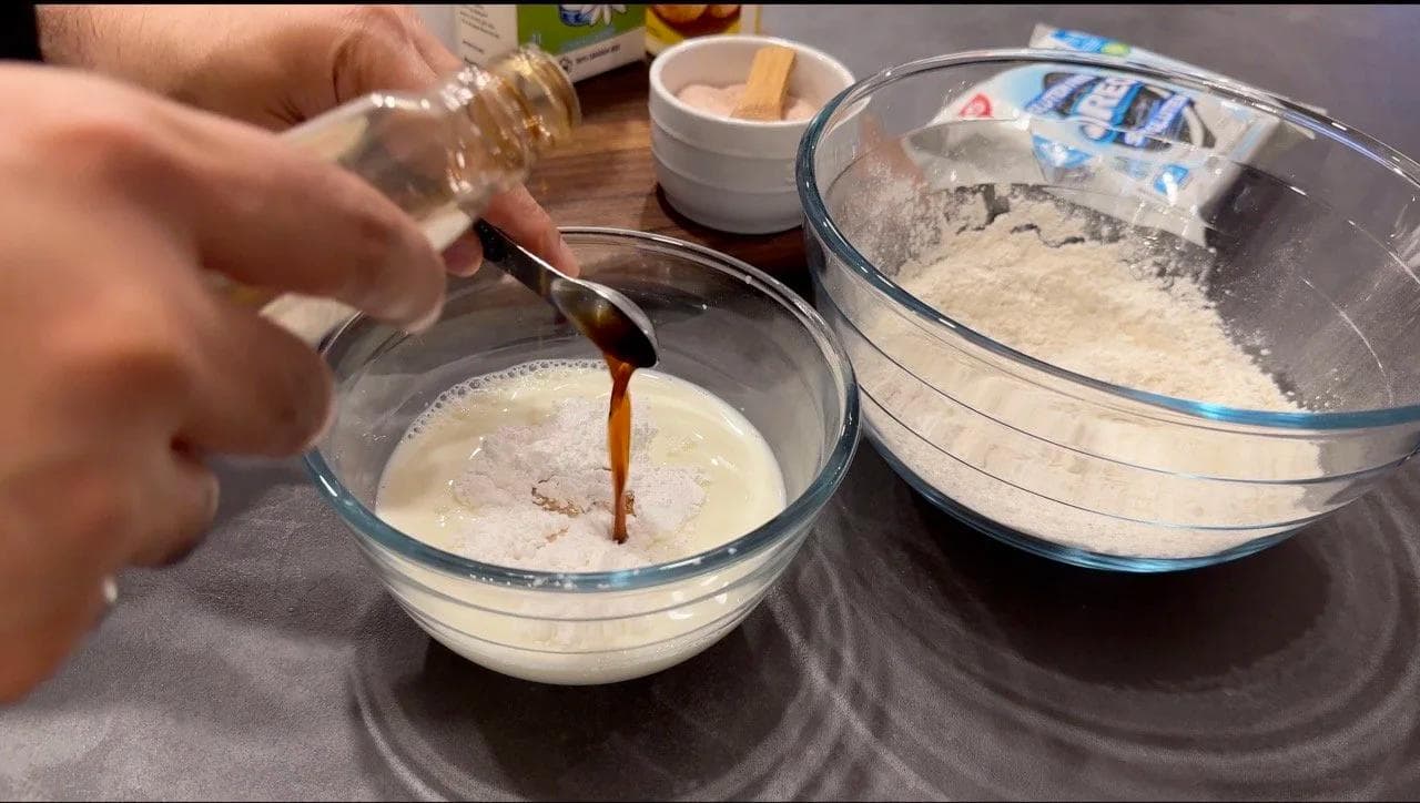 Pouring vanilla extract into a bowl of wet pancake batter next to dry ingredients in another bowl.