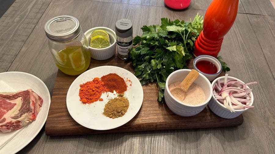 Ingredients for Brazilian churrasco laid out on a wooden board including raw steak, spices, fresh cilantro, sliced onions, and marinades.
