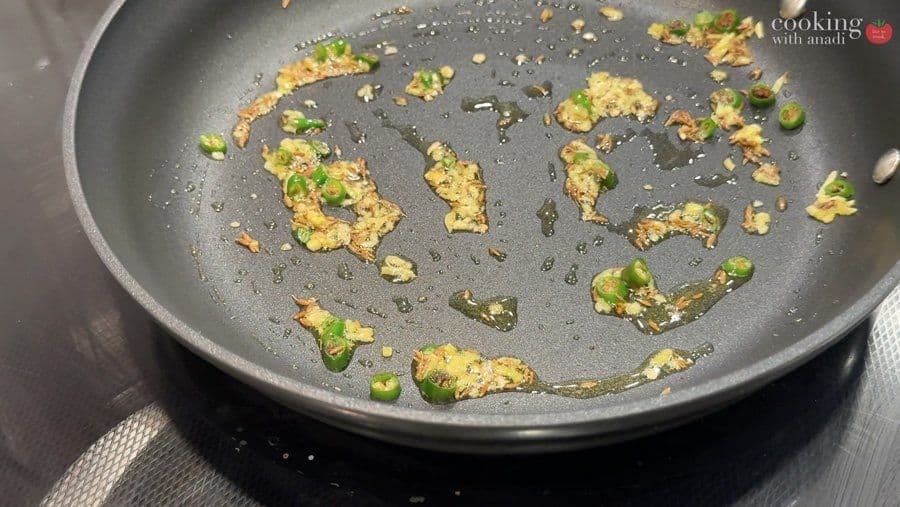 Sautéed green chiles, ginger, and cumin seeds in hot oil in a dark pan, aromatic spices cooking.