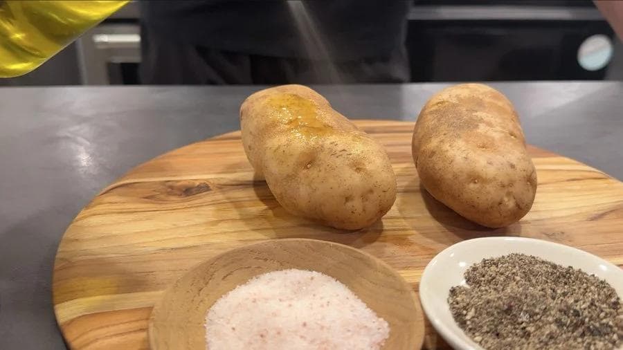 Two washed potatoes on a wooden board with bowls of salt and pepper nearby, ready for seasoning.
